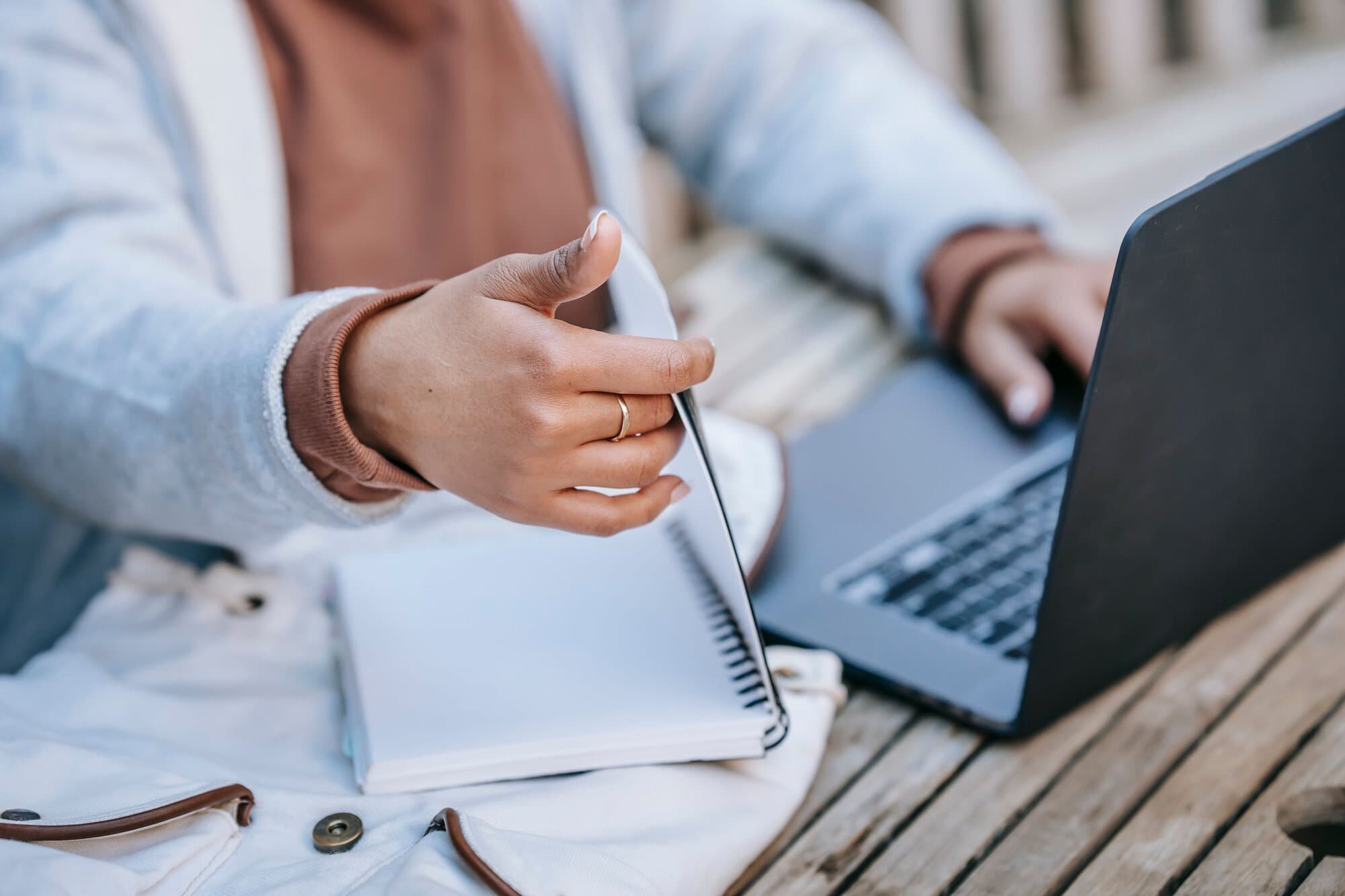 A student's hands leaf through a notebook next to an open laptop on a wooden table