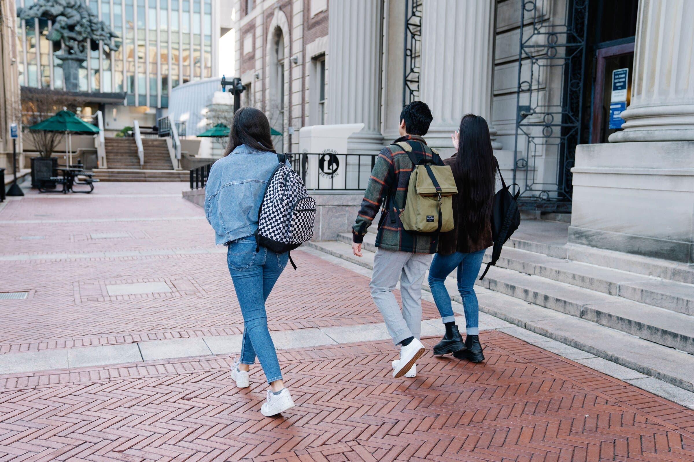 Three students with backpacks cross a brick campus plaza towards the library entrance